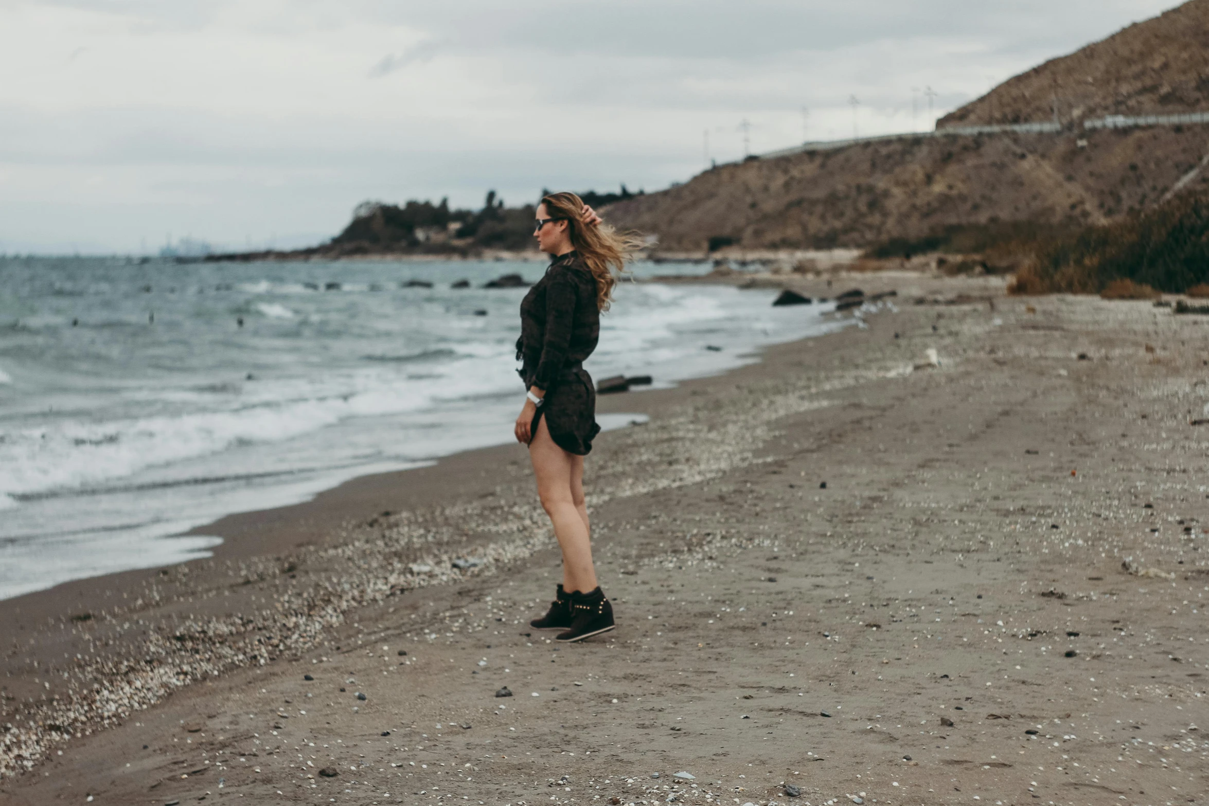 a woman standing on a beach next to the ocean, pexels contest winner, renaissance, an aviator jacket and jorts, black sea, full body image, windy day