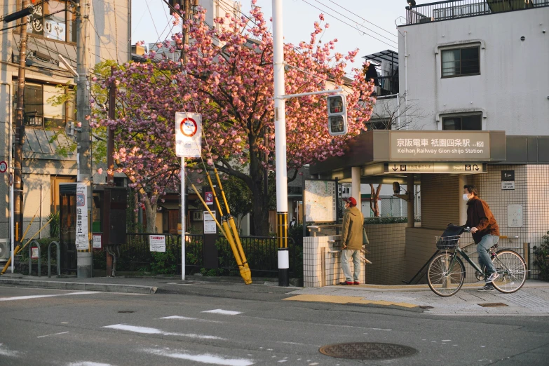 a man riding a bike down a street next to a tall building, unsplash, shin hanga, lush sakura, bus stop, 🚿🗝📝