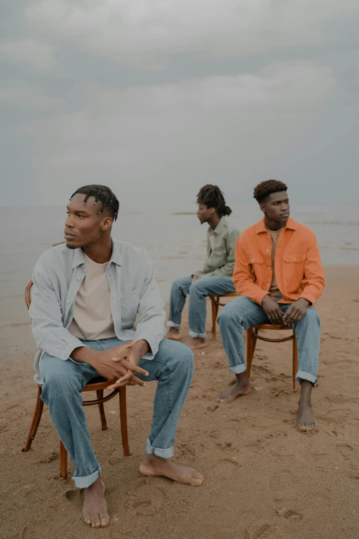 a group of men sitting on top of a sandy beach, an album cover, trending on unsplash, renaissance, wearing a jeans jackets, dark skin tone, sitting on a chair, looking serious