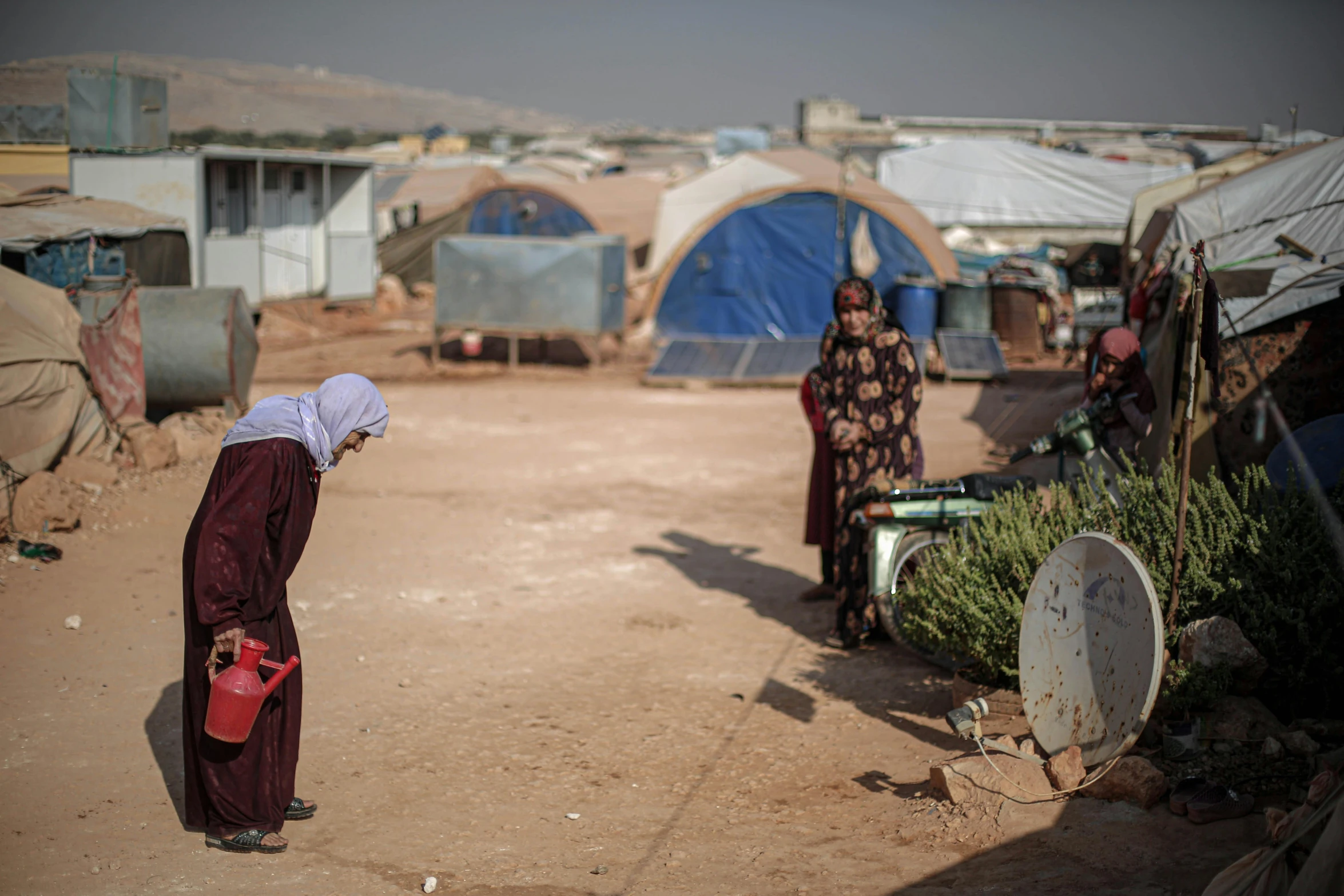 a group of women standing on top of a dirt field, hurufiyya, tent camp in foreground, white buildings with red roofs, thirst, profile image