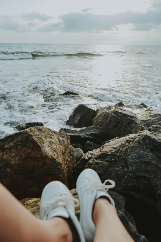 a person sitting on a rock next to the ocean, wearing white sneakers, ((rocks)), oceanside