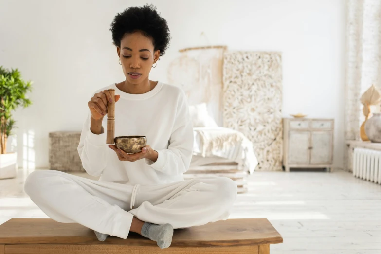 a woman sitting on top of a wooden table holding a bowl, inspired by Saneatsu Mushanokōji, trending on pexels, wearing white pajamas, holding a golden bell, wearing a tracksuit, african american woman