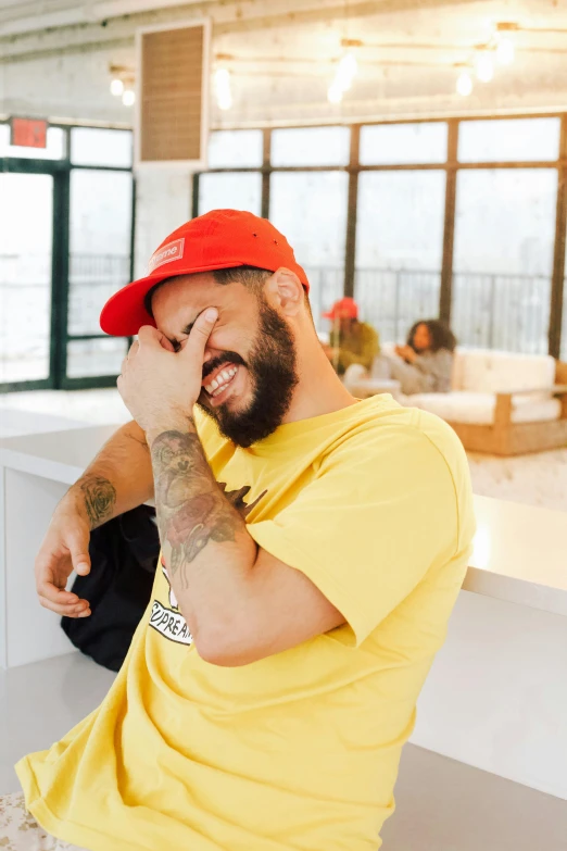 a man sitting on a bench talking on a cell phone, wearing a modern yellow tshirt, red cap, caio santos, winking