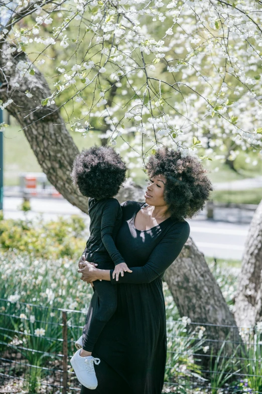 a woman holding a child in her arms, by Adam Saks, pexels contest winner, lush trees and flowers, curly afro, central park, panel of black