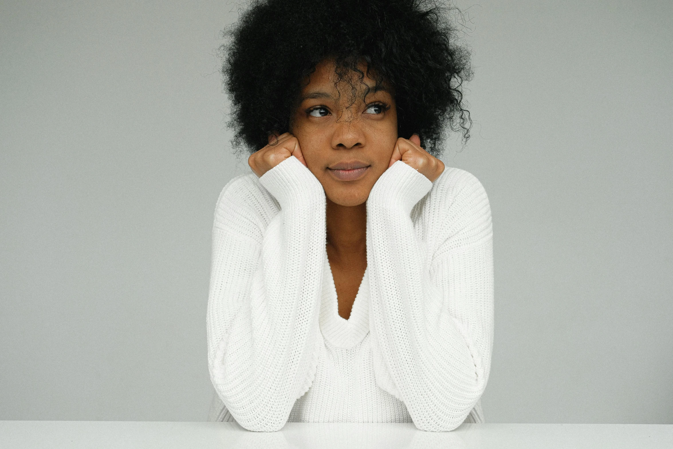 a woman sitting at a table with her hands on her face, by Lily Delissa Joseph, pexels, minimalism, wearing a white sweater, with afro, thoughtful ), various posed