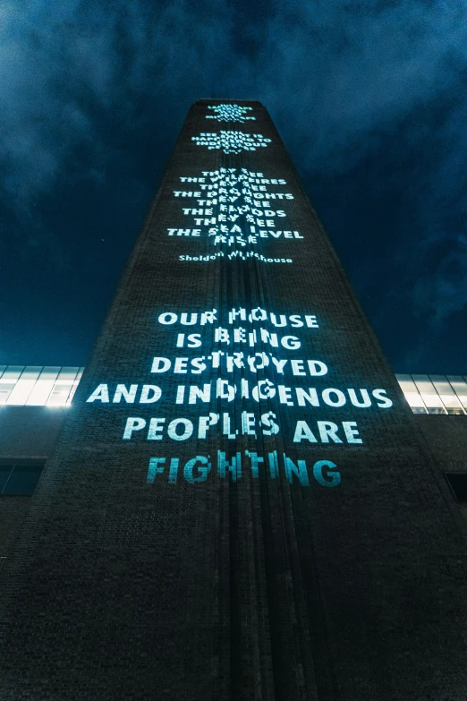 a tall building with words projected on it, inspired by Ian Hamilton Finlay, unsplash, brutalism, people fighting, obelisk, indigenous, poor lighting