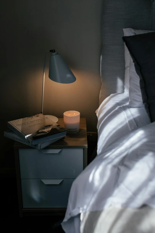 a bed room with a neatly made bed and a night stand, inspired by Eero Järnefelt, unsplash, light and space, candle lit, close up angle, books on side table, soft grey and blue natural light