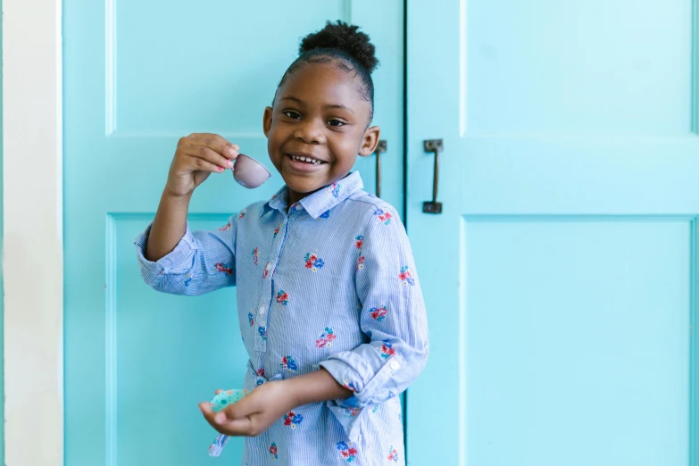 a little girl standing in front of a blue door, by Lily Delissa Joseph, pexels contest winner, holding easter eggs, earing a shirt laughing, ( ( dark skin ) ), tamagotchi