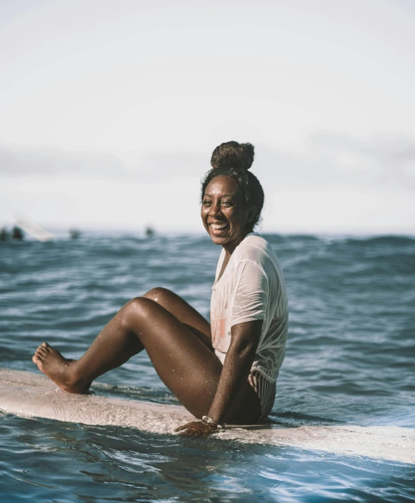 a woman sitting on a surfboard in the ocean, with brown skin, smiling, proud, iridescent skin