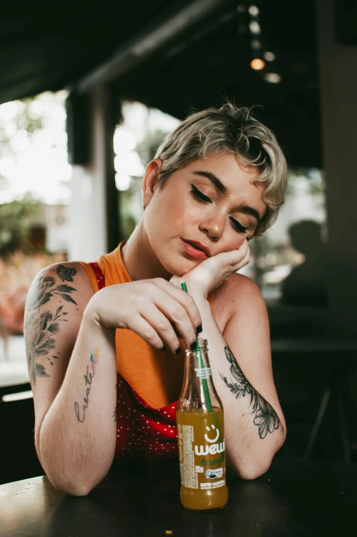 a woman sitting at a table with a bottle of beer, a tattoo, inspired by Elsa Bleda, trending on pexels, pixie cut, charli bowater, amy sol in the style of, alanis guillen