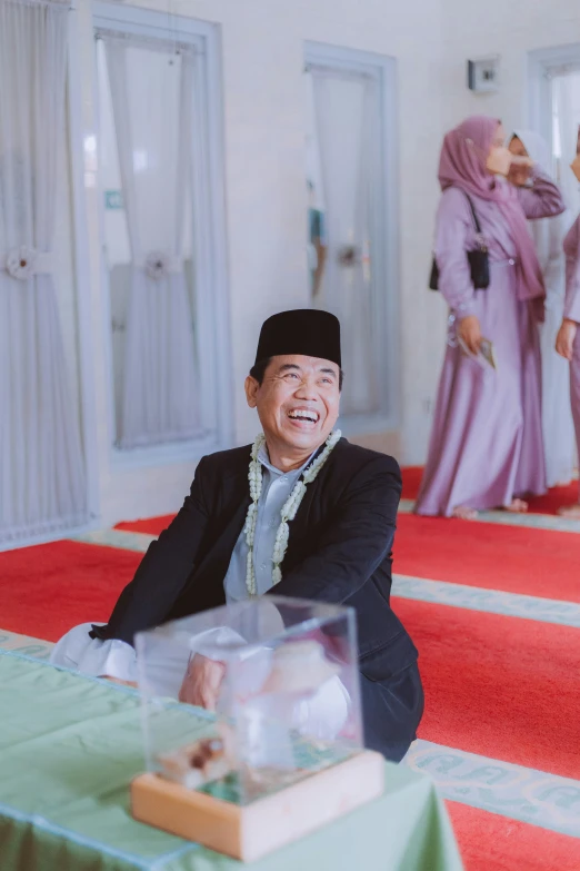 a man sitting on a red carpet in a room, by Basuki Abdullah, laughing groom, mosque, official store photo, smirking