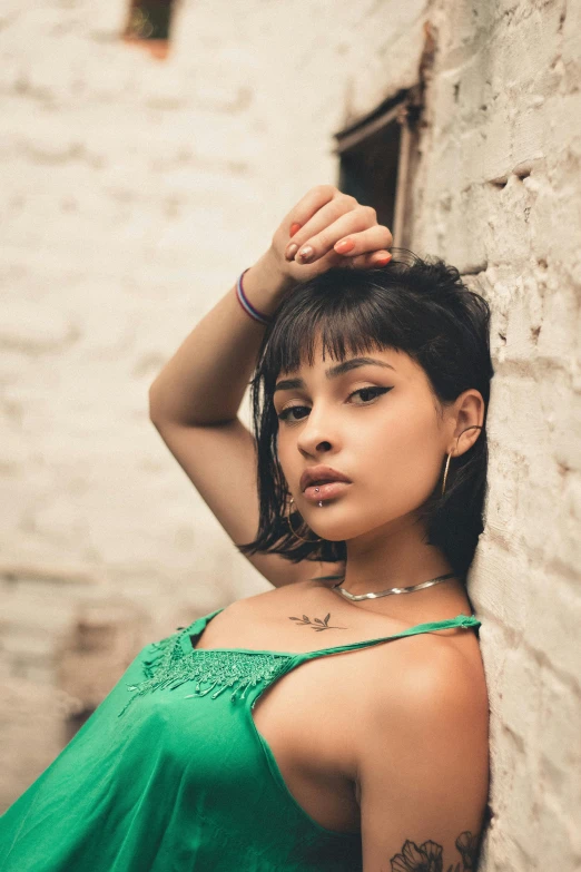 a woman in a green dress leaning against a brick wall, an album cover, inspired by Elsa Bleda, trending on pexels, isabela moner, woman in streetwear, headshot, provocative indian