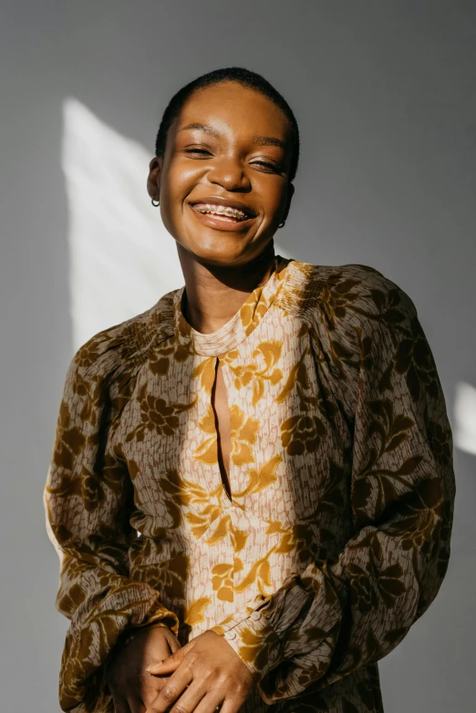 a woman standing in front of a white wall, trending on pexels, renaissance, wearing yellow floral blouse, ( ( dark skin ) ), laughing, he is wearing a brown sweater