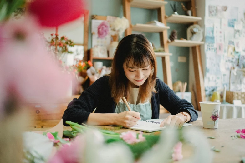 a woman sitting at a table writing on a piece of paper, a drawing, inspired by Yukimasa Ida, pexels contest winner, flower shop scene, avatar image, vietnamese woman, instagram picture