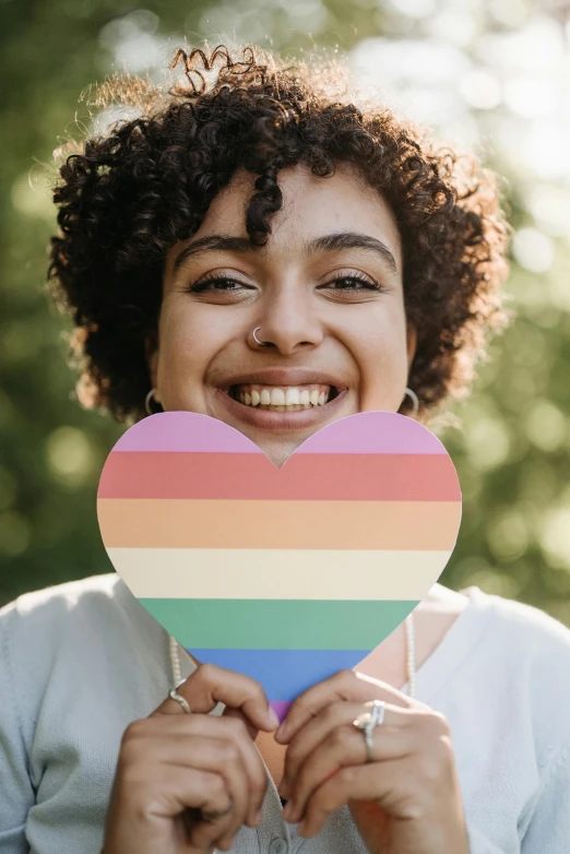 a woman holding a rainbow heart in front of her face, trending on pexels, dark short curly hair smiling, holding a wooden staff, photo of young woman, androgynous male