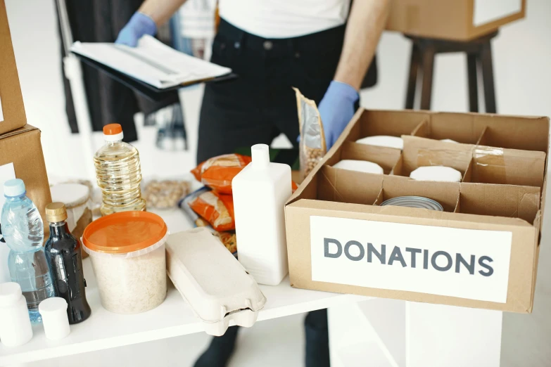 a woman holding a clipboard in front of a box of donations, by Francis Helps, pexels contest winner, on a white table, 🦩🪐🐞👩🏻🦳, emergency, tins of food on the floor