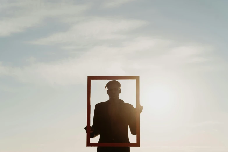 a man holding a picture frame in front of the sun, by Attila Meszlenyi, pexels contest winner, surrealism, square pictureframes, clear silhouette, natural light outside, minimalistic art