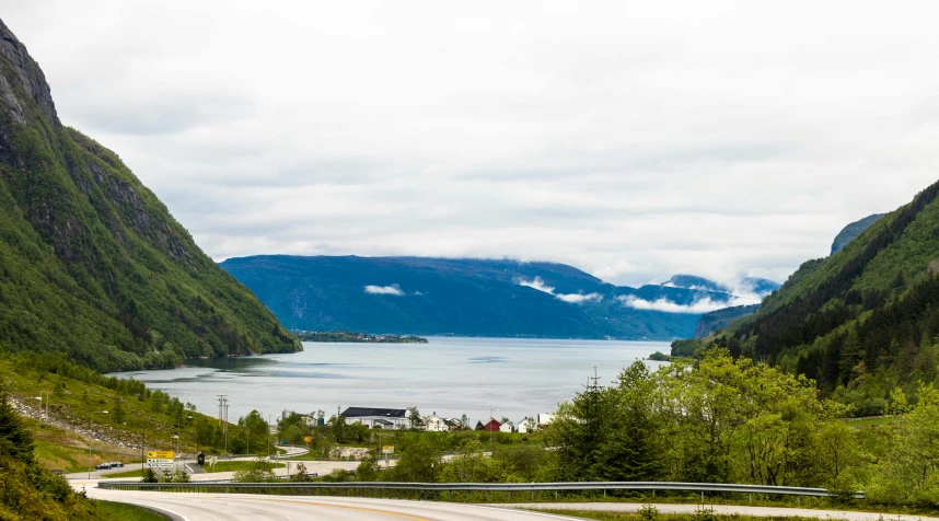 a car driving down a winding road next to a body of water, by Roar Kjernstad, distant town in valley and hills, conde nast traveler photo, festivals, panoramic