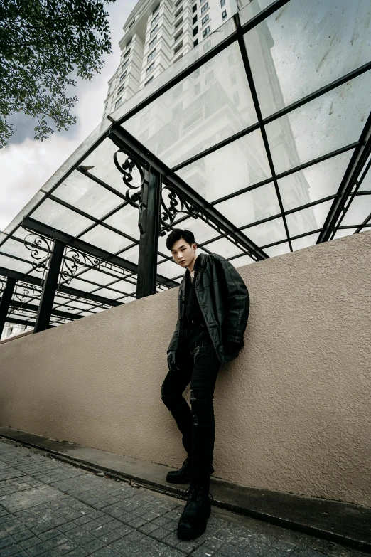 a man leaning against a wall in front of a building, an album cover, inspired by Zhang Han, unsplash, bauhaus, black leather jacket, elegant pose, rooftop, set on singaporean aesthetic