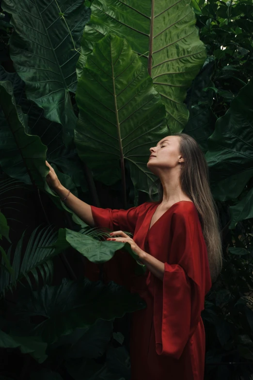 a woman in a red dress standing in front of large green leaves, by Emma Andijewska, pexels contest winner, renaissance, gently caressing earth, dance meditation, tall plants, long hair and red shirt