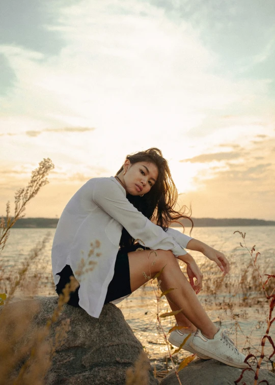 a woman sitting on top of a rock next to a body of water, trending on pexels, romanticism, wearing a white button up shirt, goldenhour, sad look, concert