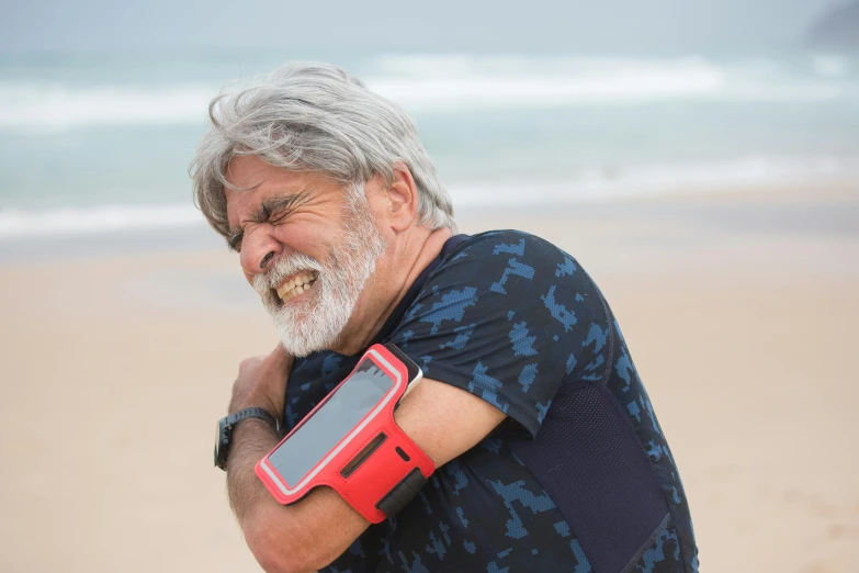 a man standing on top of a sandy beach next to the ocean, huge shoulder pauldrons, in pain, he is holding a smartphone, old gigachad with grey beard