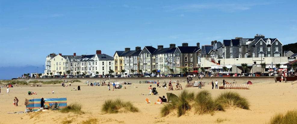 a group of people standing on top of a sandy beach, victorian buildings, relaxing on the beach, wales, a cozy