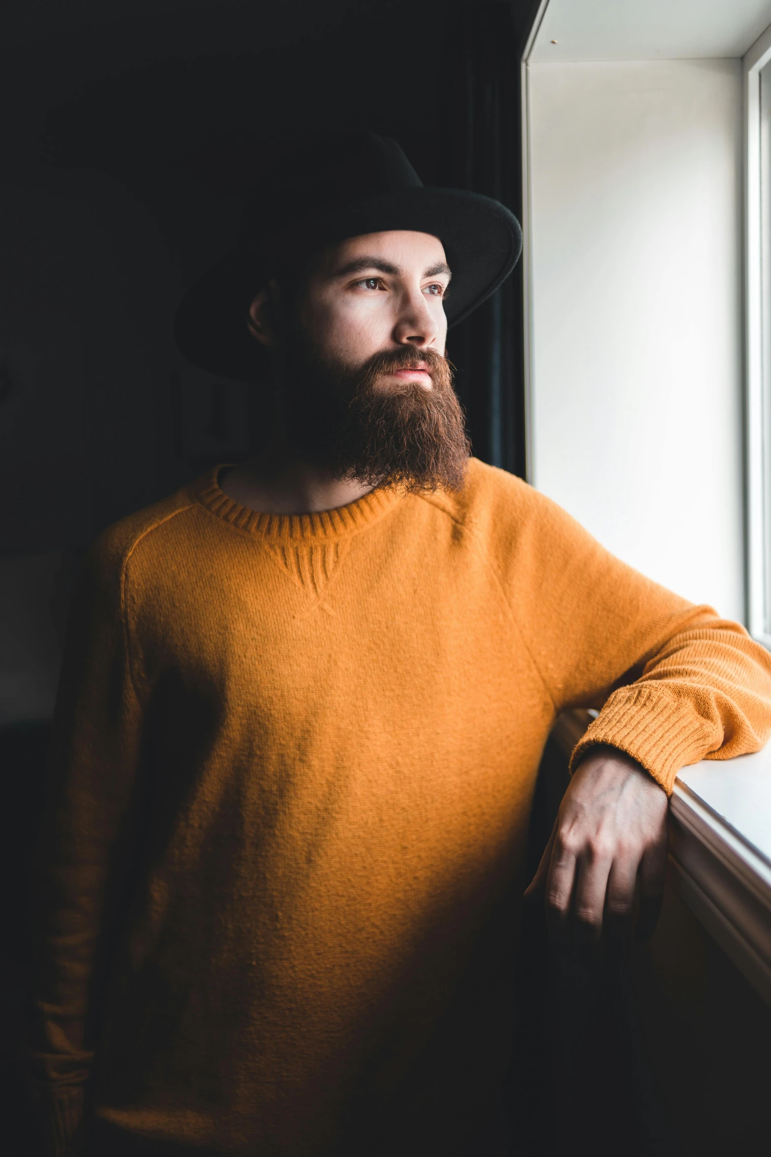 a man with a beard standing in front of a window, pexels contest winner, long orange sweatshirt, a suited man in a hat, lachlan bailey, avatar image