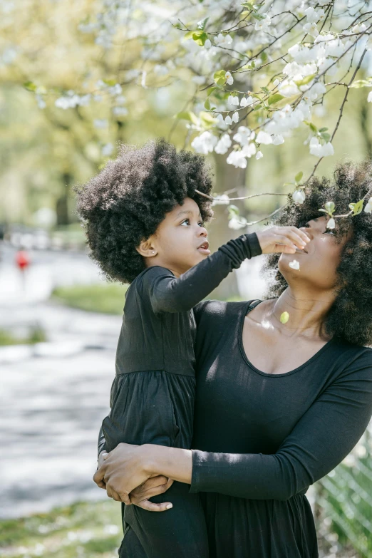 a woman holding a child in her arms, by Lily Delissa Joseph, pexels contest winner, detailed trees in bloom, black curly hair, picking up a flower, fashionable