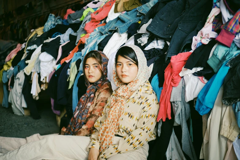 two women sitting next to each other in front of a pile of clothes, a colorized photo, unsplash, hurufiyya, in a warehouse, wearing a head scarf, sana takeda, ignant