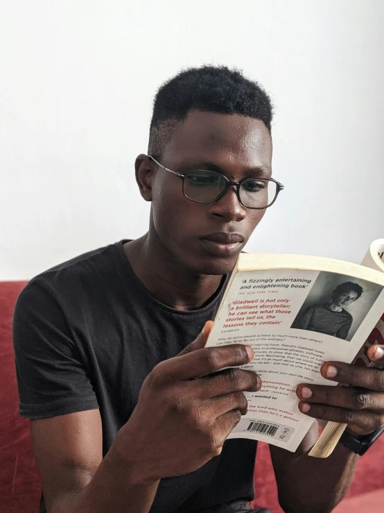 a man sitting on a couch reading a book, by Lily Delissa Joseph, pexels contest winner, afrofuturism, franz schubert look alike, 21 years old, wearing black frame glasses, profile picture