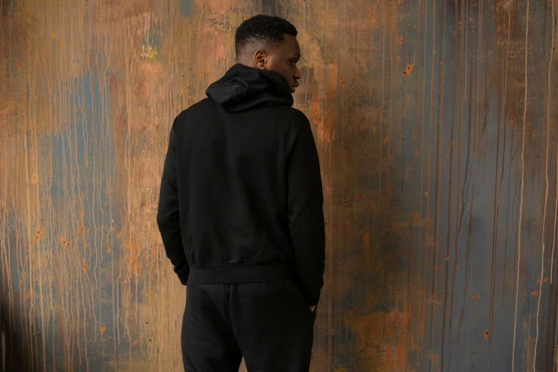 a man standing in front of a rusty wall, inspired by Paul Georges, dressed black hoodie, seen from the side, wearing a track suit, vantablack cloth technology