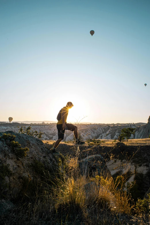 a person standing on a rock with hot air balloons in the background, at the golden hour, running freely, combat and adventure photography, stretch