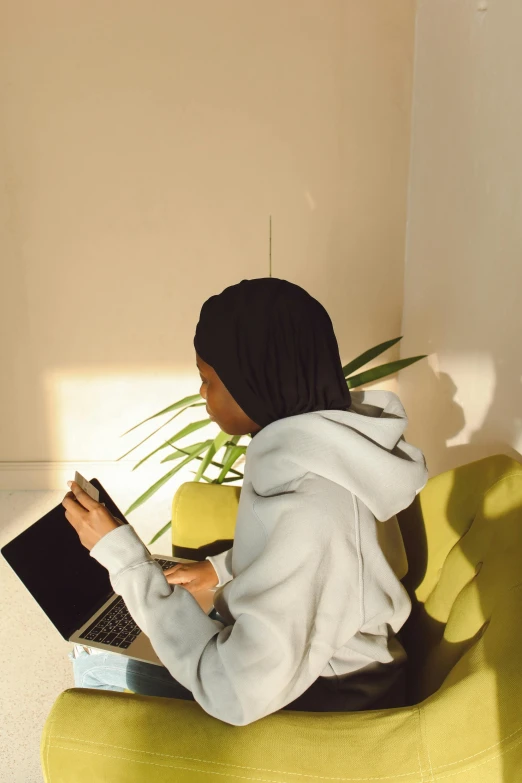 a woman sitting on a yellow chair with a laptop, by Lily Delissa Joseph, trending on pexels, realism, black haired girl wearing hoodie, next to a plant, back light, holding notebook