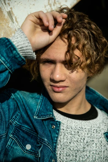 a young man in a denim jacket leaning against a wall, an album cover, by Jacob Toorenvliet, trending on pexels, curly blond hair, portrait close up of guy, attractive androgynous humanoid, brown haired