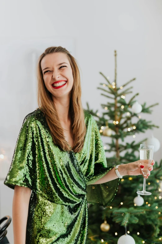 a woman in a green dress holding a glass of wine, wearing festive clothing, sequin top, with a tall tree, smiling