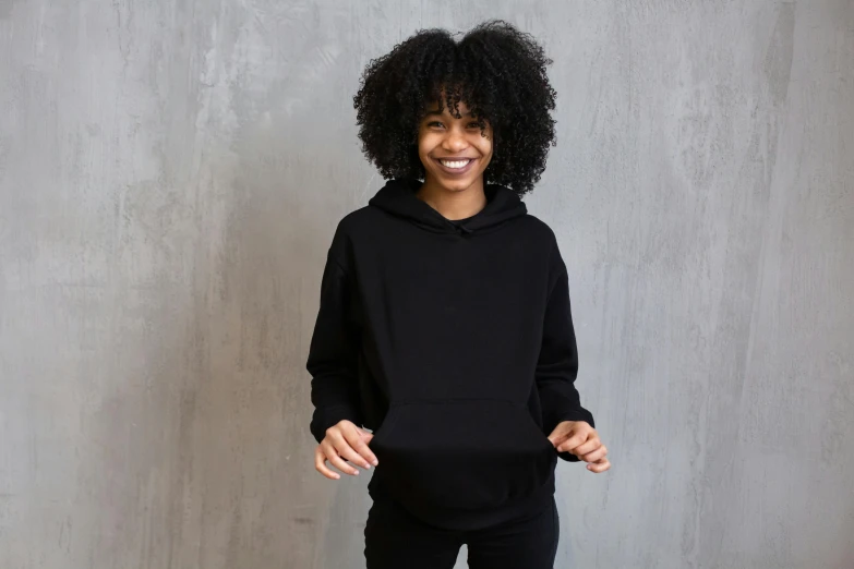 a woman standing in front of a wall wearing a black hoodie, dark short curly hair smiling, plain background, no - text no - logo, hands on hips