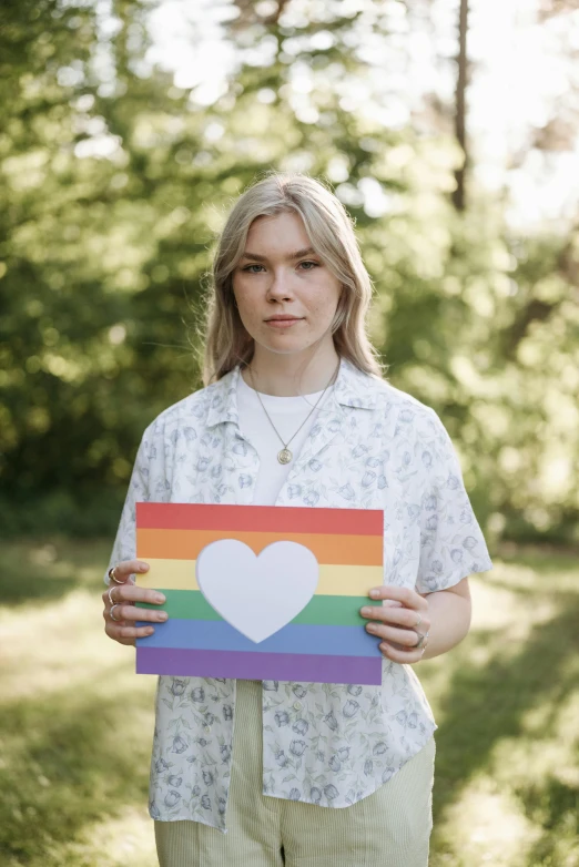 a woman holding a rainbow heart sign in a park, by Julia Pishtar, kalevala, 2 4 - year - old man, profile image, patriotism