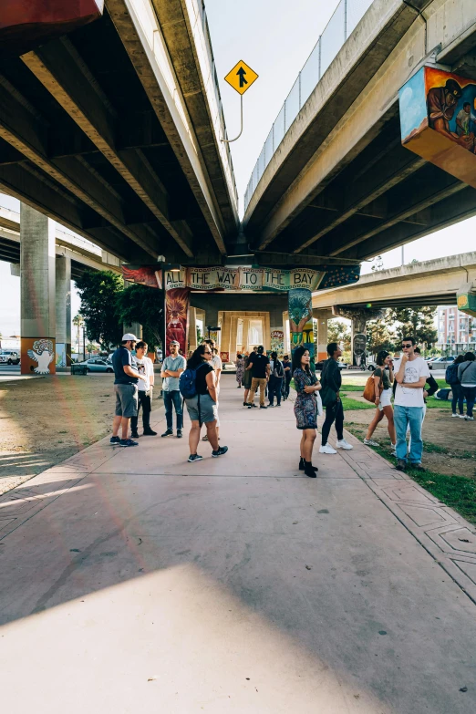 a group of people standing on a sidewalk under a bridge, by Alejandro Obregón, unsplash, happening, public art, very wide view, los angelos, instagram story