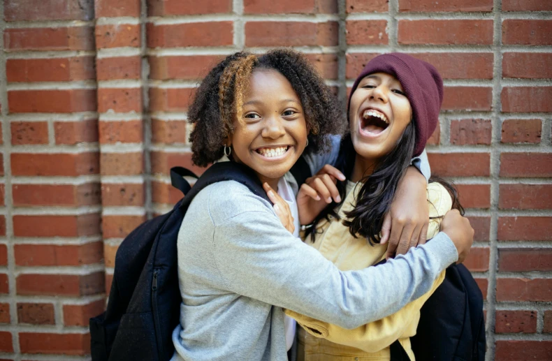 two girls hugging each other in front of a brick wall, trending on pexels, happening, black teenage girl, welcoming grin, background image, a backpack