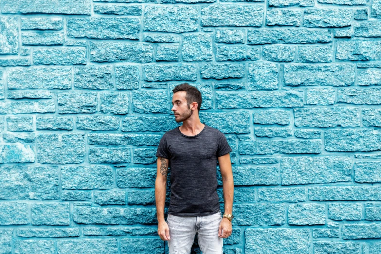 a man standing in front of a blue brick wall, by Kristian Zahrtmann, pexels contest winner, wearing a black tshirt, 9 9 designs, street of teal stone, background image