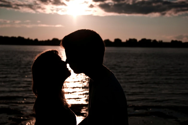 a couple standing next to each other in front of a body of water, by Carey Morris, pexels contest winner, romanticism, silhouette :7, kissing together cutely, mega-detailed, late summer evening