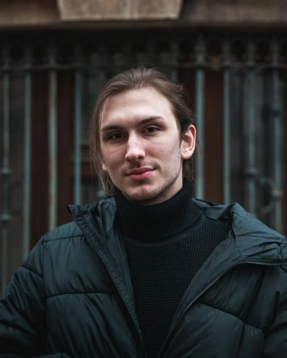 a man in a black jacket standing in front of a building, inspired by Andrei Ryabushkin, trending on unsplash, nonbinary model, lovingly looking at camera, wearing turtleneck, russian academic