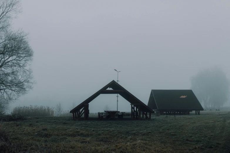 a couple of tents sitting on top of a grass covered field, by Attila Meszlenyi, pexels contest winner, postminimalism, next to a farm house and a barn, eerie fog, wooden structures, grey