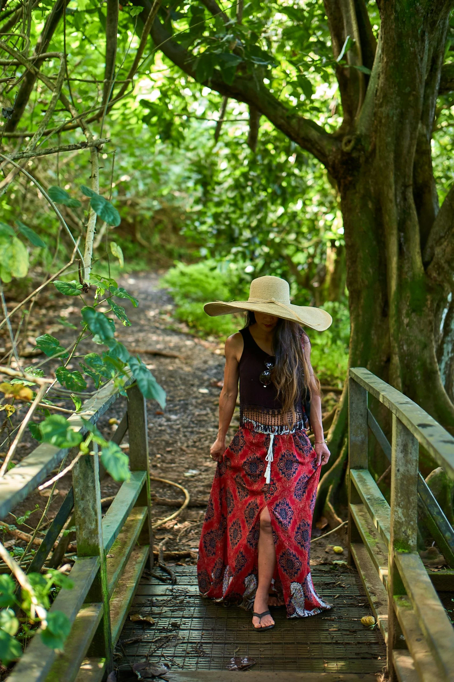 a woman in a hat is walking on a bridge, by Alice Mason, unsplash, in the tropical wood, cornwall, tribal clothing, on a green hill