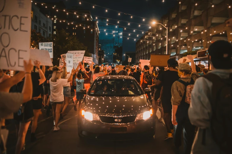 a car driving down a street next to a crowd of people, by Benjamin Block, pexels contest winner, renaissance, protest, lights on, cardboard, : :