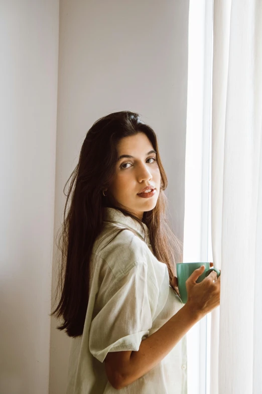 a woman standing in front of a window holding a cup, inspired by Elsa Bleda, trending on pexels, renaissance, mahira khan as a mage, long coffee brown hair, sea - green and white clothes, portrait image