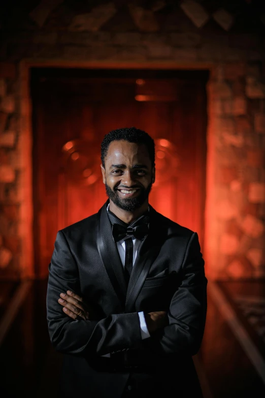 a man in a tuxedo standing in front of a red door, inspired by Edwin Georgi, on a dark rock background, dark-skinned, with a beard and a black shirt, looking happy