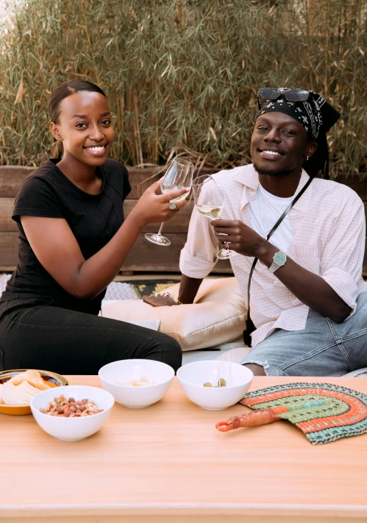 a couple of women sitting on top of a wooden table, very kenyan, holding glass of wine, humus, riyahd cassiem