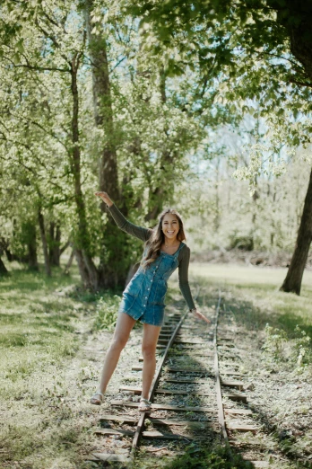 a woman in a blue dress standing on a train track, a picture, by Kristin Nelson, unsplash, happening, standing in grassy field, fun pose, walking on an old wood bridge, high school